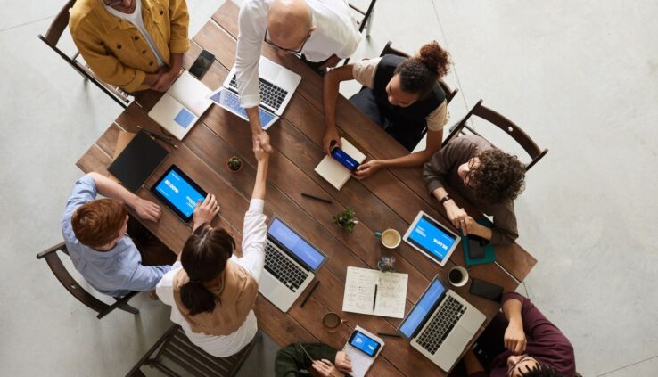 Overhead view of a diverse team around a wooden table with laptops, tablets showing 85 percent, and a handshake