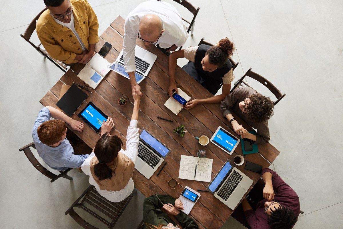 Overhead view of a diverse team around a wooden table with laptops, tablets showing 85 percent, and a handshake